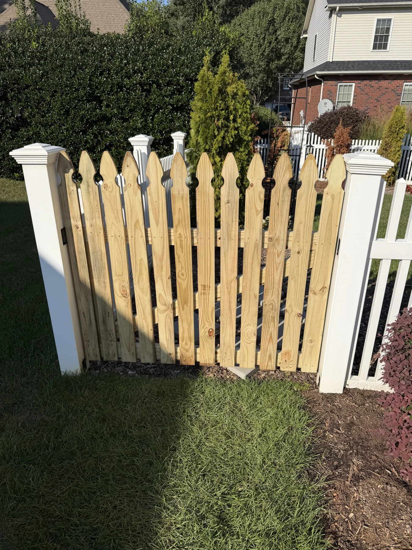 A custom wood picket gate fitted between existing posts. Front and angled views show the shaped pickets, frame and connection to the adjoining fence.
