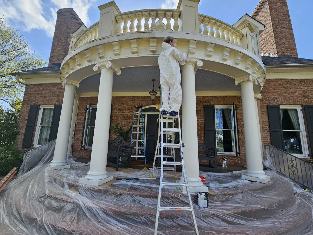 An exterior carpentry work area at a curved, columned porch with an upper balustrade. The photograph shows protective coverings and access equipment while the crew works on the architectural details.