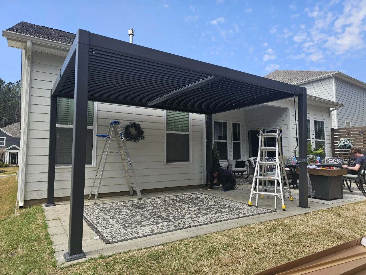 An aluminum pergola installation adds a defined outdoor gathering area alongside the home. These progress photographs show the structure and its clean architectural lines.